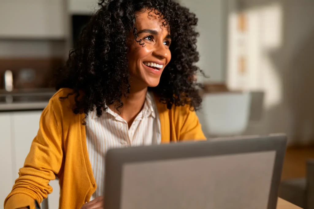 Mulher sorridente de cabelos cacheados trabalha em um laptop, sentada à mesa em uma cozinha iluminada.
